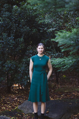 A woman stands amidst dense greenery, her confident pose and a hint of a smile blending beautifully with the vibrant natural surroundings and sunlight