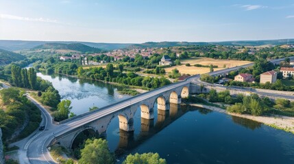 Fototapeta premium River crosses under old stone bridge, rural town in background.