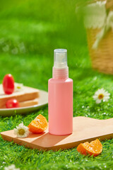 In the center of a wooden tray, surrounded by a tomato sandwich, orange slides, and a rattan basket, is a frontal view of a mockup of a pink spray bottle. A picnic-themed image in lush grass.