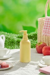 A template image with a picnic landscape theme, a white carpet with a tea set, macaroons, apples, books, baskets, and an empty label pump bottle in yellow color, on green grass. Frontal view.