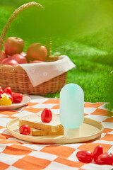 A sunscreen advertisement photo with a picnic concept, a front of an unbranded blue bottle placed on a ceramic tray, next to breads, tomatoes, and an apple basket scattered on a mat. Grass backdrop.