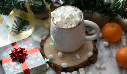 White cup of a hot chocolate with marshmallows on top, Christmas decorations and a Christmas tree, mandarins and cookies as a background. Warm Holidays season