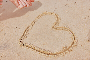 Child's hand drawing heart shape in sand on beach