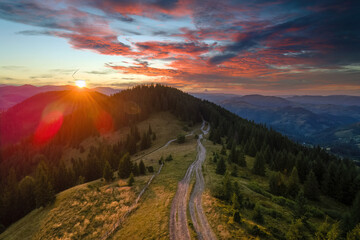 Beautiful nature landscape. Aerial view of colorful sunset in wild mountains. Dark dirt road illuminated with bright setting sun