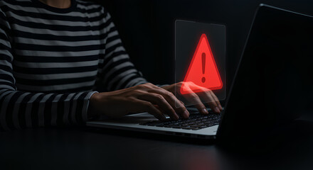 A person typing on a laptop with a red warning triangle displayed on the screen.