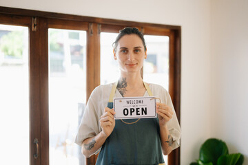 Young female entrepreneur hanging a welcome sign in front of a coffee shop. Beautiful waitress or...