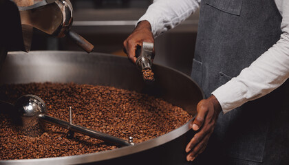 African male barista in uniform apron testing aroma of fresh hot coffee beans in roasting manufacturing.