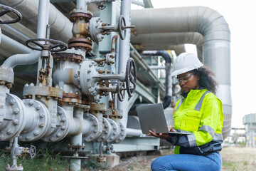 A female engineer wearing a safety helmet and high-visibility jacket works on a laptop beside an industrial pipeline system,highlighting engineering,technology, and maintenance in a complex industrial