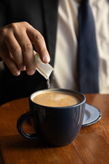 Unrecognizable businessman in suit working and pouring a sugar packet into a steaming cup of coffee where the sugar grains are falling in close up on his wooden desk in his office
