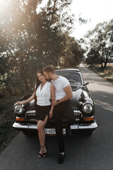 couple standing in front of a classic black car parked on the road. A man in a white T-shirt and black trousers hugs a woman