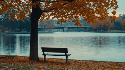 Obraz premium Serene autumn scene empty park bench by lake under tree with fall leaves, bridge in background.