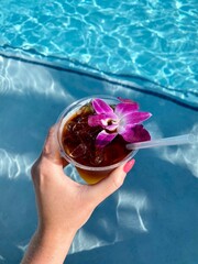 Hand holding a refreshing beverage garnished with an orchid by a sparkling blue poolside on a sunny day