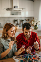 Loving couple collaborate and smile to create beautiful Easter egg designs at home.