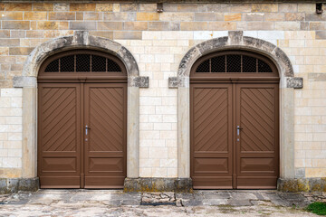 Two entrances, two brown wooden doors against a stone block wall. From the series of doors of the World.