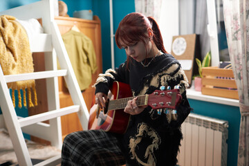 Medium shot of modern gen Z Caucasian girl standing in cluttered dorm room, playing guitar