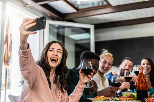 Group of cheerful friends taking a selfie while enjoying wine and food together. Multicultural gathering with laughter and celebration in a bright and relaxed indoor setting.