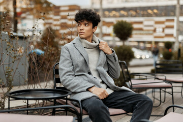 Fashionable young man in grey coat sitting on bench with coffee cup in hand at outdoor table