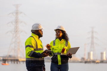 Two engineers in safety helmets and high-visibility jackets discuss work while holding a tablet and...