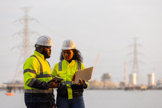 Two engineers in safety helmets and high-visibility jackets discuss work while holding a tablet and clipboard at a power plant site,with transmission towers and industrial structures in the background