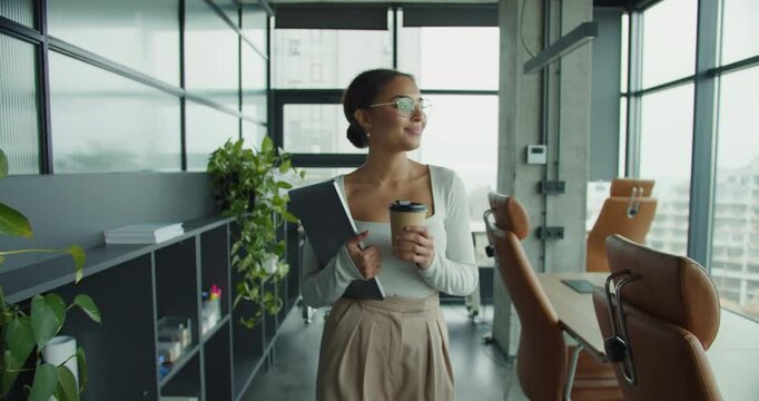 young professional woman with glasses and coffee cup walking confidently in a modern office, holding a laptop and smiling while looking out large glass windows with natural lighting