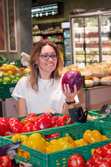 A woman in her 30s-40s selects fresh fruits and vegetables in a supermarket. The photo emphasizes organic food, healthy eating, vegan lifestyle, sustainable living, and mindful nutrition choices.