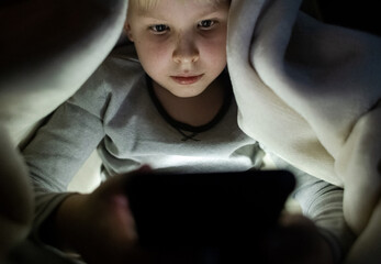 Child phone addiction. The boy at night, covered with a blanket, on the bed plays games on the smartphone and watches videos, cartoons