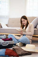 Asian women preparing clothes in suitcases. She's choosing clothes, travel documents, itinerary for a solo trip, and checking the checklist in her living room in travel holiday concept