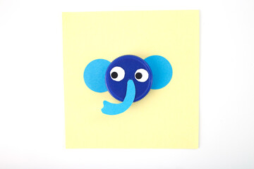 Blue plastic bottle cap transformed into elephant face with paper ears and trunk on a yellow square paper. Minimalist flat lay composition Craft and DIY recycling concept.