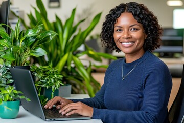 Smiling African woman in a blue jumper working on her laptop in a plant-filled workspace. Copy space.