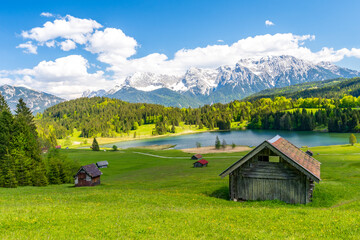 A stunning view of Geroldsee lake, framed by wooden huts and the snow-covered Karwendel mountains against a vibrant blue summer sky in Bavaria, Germany