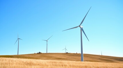 Fototapeta premium Wind turbines stand tall on a golden hillside, harnessing renewable energy under a clear blue sky.