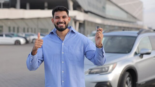 Attractive male driver pointing at keys from new car. Portrait of smiling dark-haired man showing keys to new car with ok gesture, gesturing great choice. Concept of emotions, purchase, happiness.