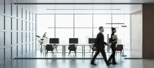 Modern office workspace with glass walls, desks, and computers. Two businesspeople walking past. Bright city view in the background.
