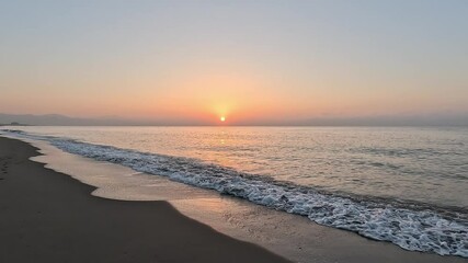 Handheld video of the sun at sunrise or sunset over the horizon of the calm Mediterranean Sea, with clear sky at golden hour, seen from the beach with the waves coming over the sand