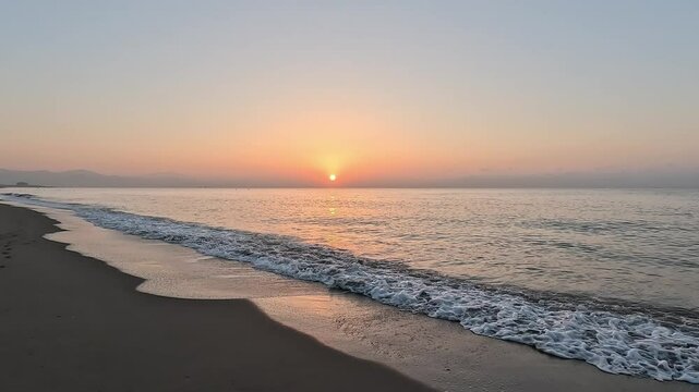 Handheld video of the sun at sunrise or sunset over the horizon of the calm Mediterranean Sea, with clear sky at golden hour, seen from the beach with the waves coming over the sand