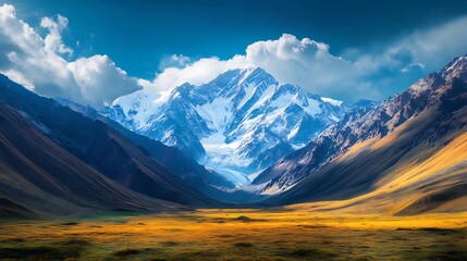 Majestic snow-capped mountain peak dominates a valley with golden grasslands under a vibrant blue sky.
