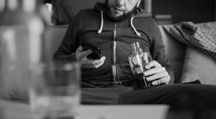 man sitting on sofa home holds smartphone in one hand and bottle of alcohol in the other. Stress. Loneliness