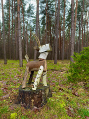A forest scene in autumn, featuring a tree stump surrounded by pine trees and a meadow. In the foreground, a deer made from firewood, complete with antlers, stands proudly on the stump. The image capt