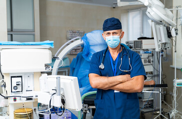 Naklejka premium Health pro in a modern hospital. A male doctor stands confidently in a hospital room, wearing scrubs and a mask, surrounded by medical equipment.