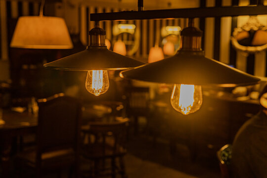 Two pendant lamps in a dimly lit room hanging from a black metal fixture. Night cafe interior. Warm light. Defocused interior of restaurant or bar, with tables and chairs visible.