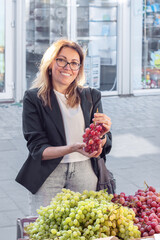 A woman in her 30s-40s selects fresh grapes at an outdoor market. The photo highlights organic...