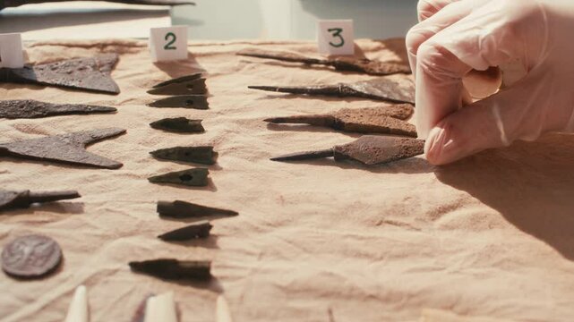 Gloved hand of unrecognizable archaeologist placing ancient metal artifacts on table covered with soft cloth, with numbered labels indicating arrangement for study and categorization. Close-up view