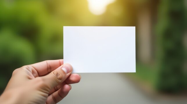 A hand holds a blank white card against a blurred natural background, offering space for custom text or design