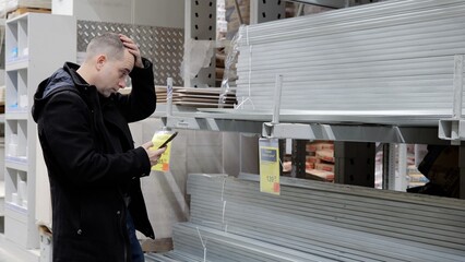 Confused male comparing smartphone prices with hardware store tags, contemplating purchasing...