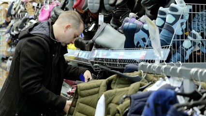 Shopper examining warm vests, checking labels in clothing store with footwear displays visible on surrounding shelves