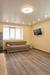 Apartment interior. Brown sofa with bedside table in the living room. TV on the wall. Furniture in the living area. Linoleum on the floor. Chandelier on the ceiling.