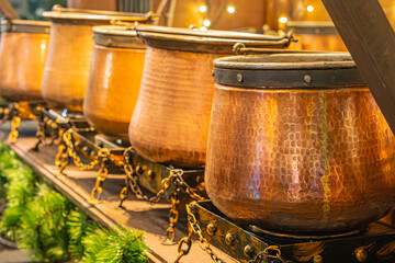 Image shows two rows of copper pots with lids, arranged on a metal rack, reflecting light Background has decorative elements, possibly taken in a kitchen or display setting for promotional use