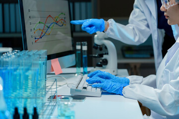 Two scientists wearing lab coats and gloves are analyzing data displayed on a computer screen in a laboratory, with test tubes and a microscope nearby