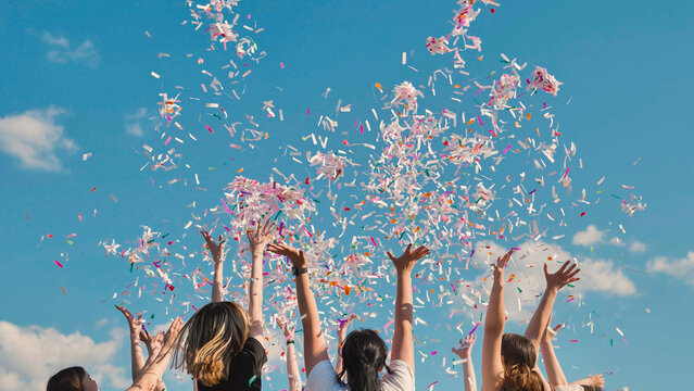 Friends celebrating joyfully outdoors, throwing colorful confetti under a blue sky. Arms raised in excitement, surrounded by a cheerful atmosphere