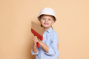 Little girl in hard hat with putty knife on beige background. Dreaming about future profession